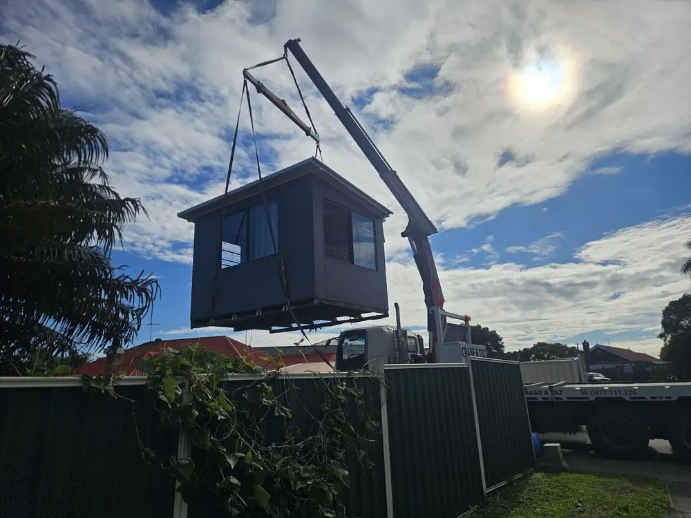 Crane truck lifting a small prefabricated structure above a fence